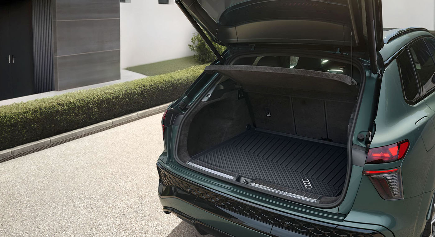 Audi Q3 SUV with an open trunk showcases a spacious, clean cargo area with a protective mat. The setting is a modern, sunlit driveway, conveying readiness and functionality.
